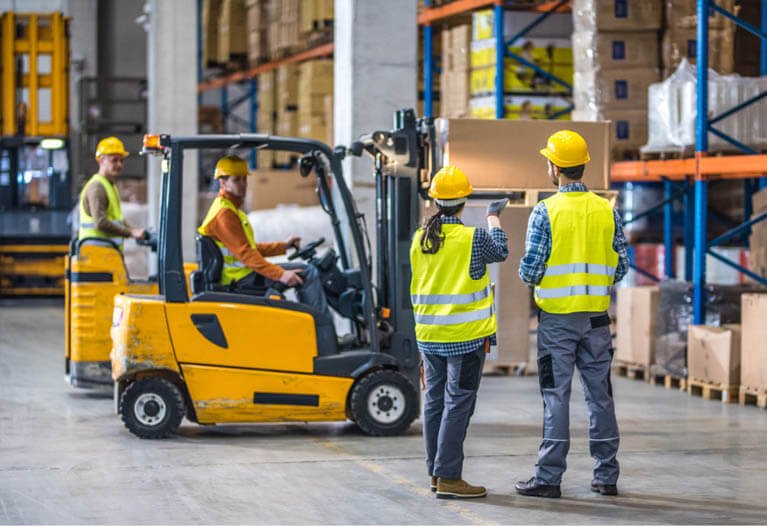 A logistics worker wearing a high-visibility vest inspects inventory in a well-lit warehouse aisle, demonstrating the need for safety apparel.